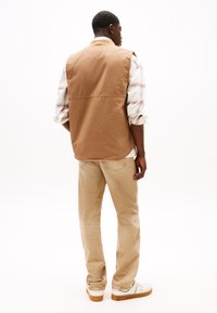 Bruine mouwloze vest over een geruit shirt, beige broek en witte sneakers. Gelaagd stof met een casual pasvorm, gezien vanuit de achterkant.