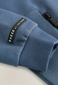 Tessuto di felpa blu navy con una finitura testurizzata, dotato di un'etichetta nera con la scritta "BAKER by TED BAKER" sul polsino e un dettaglio con zip.