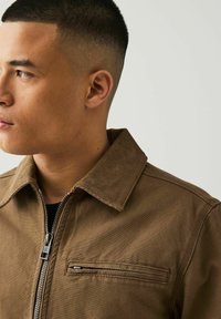 Bruine corduroy jas met een ritssluiting aan de voorkant, voorzien van een knoopsluiting en een horizontale borstzak. Glad oppervlak met subtiele stikseldetails.