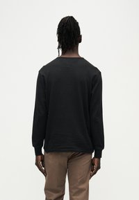 Μαύρο πουκάμισο με μακρύ μανίκι και υφή, λαιμόκοψη τύπου crew και άνετη εφαρμογή, σε συνδυασμό με καφέ παντελόνι. Το πουκάμισο δεν έχει λογότυπα ή σχέδια.