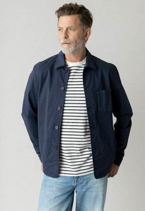 Mand med gråt skæg iført navyblå jakke med knapper over en hvid og navystribet skjorte og lyseblå jeans, der kigger til siden.