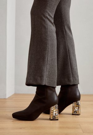 Pieds portant des bottines marron à talons blocs décoratifs et un pantalon marron à motifs, debout sur un sol en bois.