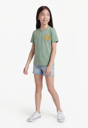 Ung jente med langt mørkt hår, iført en grønn t-skjorte med gul lapp, lyse denimshorts og hvite sneakers, står og smiler.