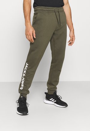Mann trägt olivgrüne Jack & Jones Sweatpants und schwarze Adidas-Sneaker, steht vor einem schlichten hellen Hintergrund.