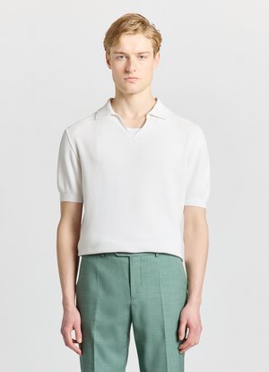 Giovane uomo in piedi, dritto, indossa una polo bianca a maniche corte con texture e pantaloni su misura verde chiaro su sfondo neutro.