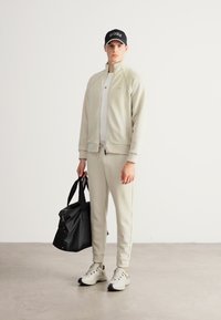 Jasnoszary dres z bluzą na zamek i zwężanymi spodniami. Czarna czapka z logo, trzyma czarną torbę podróżną i ma na sobie białe tenisówki.