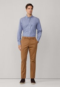 Man in een blauw geruit overhemd met knopen, in zijn bruine broek gestoken, met zwarte schoenen, staand voor een effen lichtgrijze achtergrond.