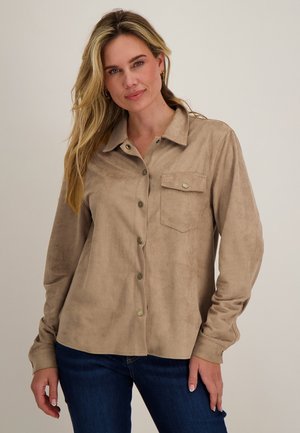 Blonde vrouw die een tan suède overhemd met knopen draagt, voorzien van een voorzak, en donkere blauwe jeans, staand tegen een effen lichte achtergrond.