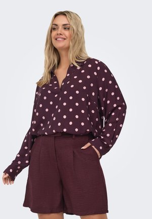 Donna che indossa una blusa a maniche lunghe viola scuro con pois rosa chiaro e pantaloncini a vita alta coordinati e dalla tessitura strutturata, sorridente e in posa.