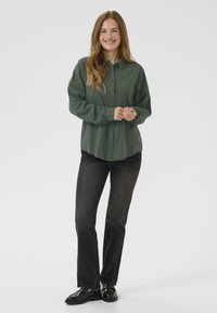 Tmavozelená košeľa na gombíky s voľným strihom, s golierom a dlhými rukávmi. Nosí sa s čiernymi džínsami a čiernymi plochými topánkami. Neutrálne pozadie.