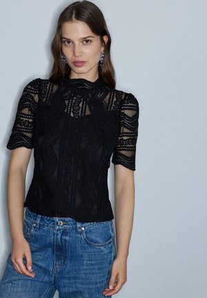 Mujer con cabello largo y castaño, vestida con un top corto de encaje negro y vaqueros azules, de pie frente a un fondo gris claro liso.