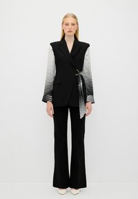 Blazer noir avec des manches à pois, cintré à la taille avec une boucle dorée. Associé à un pantalon noir évasé. Chaussures blanches.