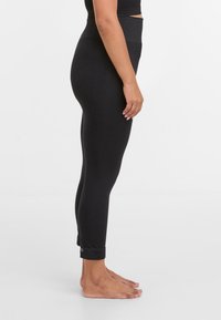 Vrouw die blootsvoets zwarte hoge taille leggings draagt tegen een eenvoudige witte achtergrond, zijaanzicht.