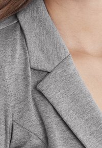 Gray blazer featuring a textured fabric, with a tailored collar and lapels. The close-up shows detailed stitching and overlapping design elements.