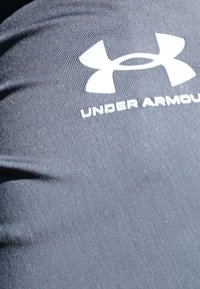 Juodas sportinis audinys su tekstūruotomis gumbeliais ir ryškia balta "Under Armour" logotipu su lygiomis raidėmis.