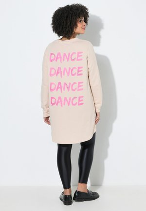 Donna con capelli ricci che indossa una felpa lunga beige con la scritta "DANCE" in lettere rosa sulla schiena, leggings neri e scarpe nere.