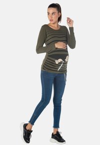 Olivengrünes Langarmoberteil mit horizontalen Streifen, das ein Cartoon-Baby-Motiv im Bauchbereich zeigt. Kombiniert mit blauen Jeans im Skinny-Style.