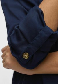 Polsino di camicia blu navy con design arrotolato, texture liscia e un bottone circolare color oro con due fori per l'inserimento.