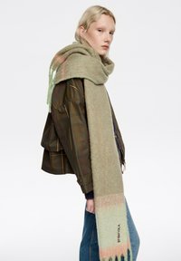 Długi, miękki szalik w pastelowej zieleni z gradientem odcieni brzoskwini, z frędzlami na krawędziach i subtelną metką marki. Noszony z ciemną kratkowaną kurtką.