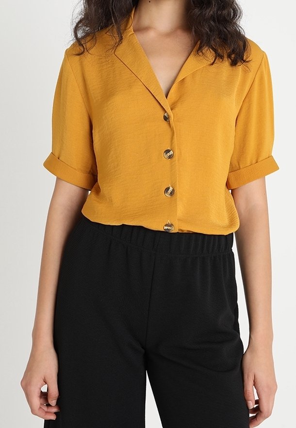 Femme portant une chemise courte à manches courtes couleur moutarde, rentrée dans un pantalon noir taille haute avec une ceinture élastique.