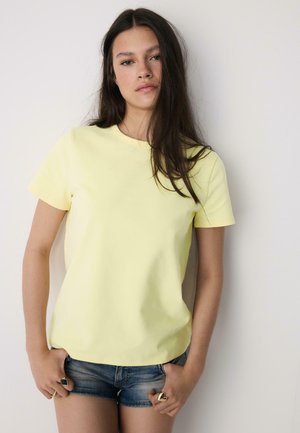 Mlada žena s dugom tamnom kosom, odjevena u svijetlo žutu majicu kratkih rukava i traper kratke hlače, stoji uz obični bijeli zid.