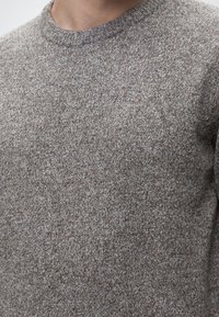 Gros plan d'une personne portant un pull en maille gris avec un petit logo Barbour brodé sur la poitrine.