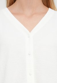 Blouse blanche boutonnée avec un col en V. Tissu texturé et cinq petits boutons circulaires sur le devant. Design simple.