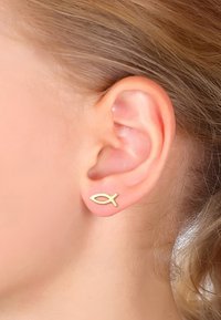 Pendiente de studs en forma de pez dorado, sujeto al lóbulo de la oreja, con textura suave, diseño minimalista, brillo sutil y apariencia ligera.