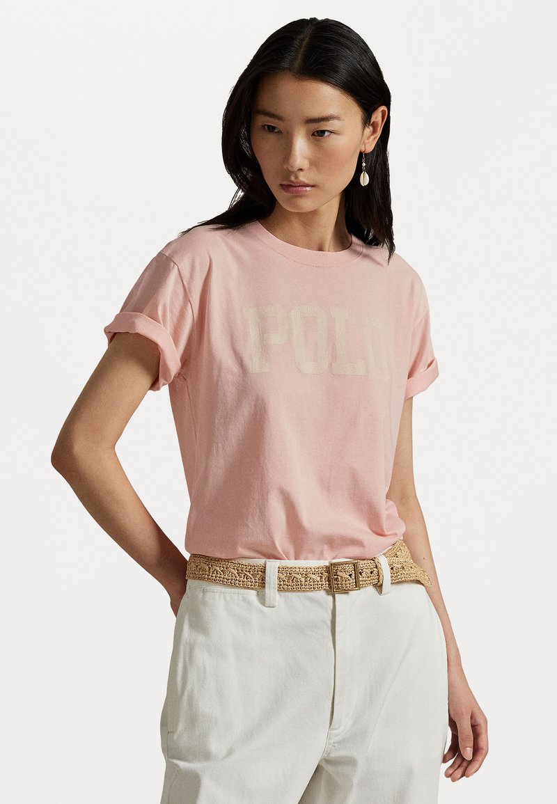 Rosa bomull t-skjorte med rullede ermer og "POLO" trykket i kremfarge. Kombinert med hvite bukser og et vevd beige belte.