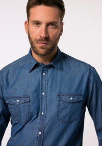 Camisa de denim en azul oscuro con cuello clásico, dos bolsillos en el pecho y cierre frontal de botones. Presenta botones plateados y una textura suave.
