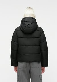 Cilvēks ar platīna blondiem matiem, ģērbies melnā kapucētā pūkainā jaku un pelēkās krāsas biksēs, redzams no aizmugures uz balta fona.