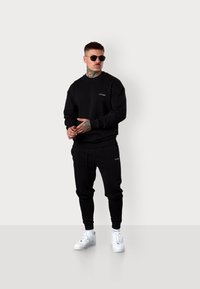 Schwarzer Pullover mit Rundhalsausschnitt und Logo auf der Brust, kombiniert mit schwarzen Jogginghosen mit Taschen. Weiße Sneakers runden das Outfit ab.