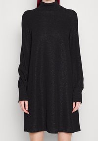 Robe noire étincelante avec un col montant, des manches longues bouffantes et une silhouette décontractée. Tissu texturé avec des paillettes, longueur mi-cuisse.