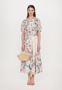 Floral blouse with short puff sleeves, paired with a floral maxi skirt. The model holds a woven cream handbag and wears nude strappy sandals.