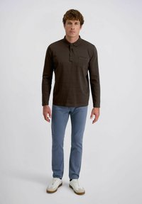 Mann in einem dunkelbraunen Langarm-Polohemd, blauen Jeans und weißen Turnschuhen, der vor einem schlichten Hintergrund steht.