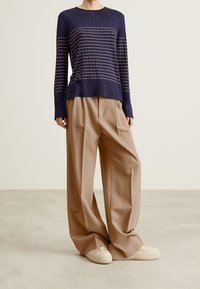 Sweter w niebiesko-białe paski z prążkowanego materiału, z długimi rękawami, w połączeniu z szerokimi beżowymi spodniami i białymi sneakersami, na neutralnym tle.