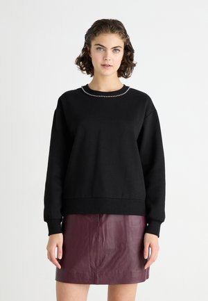 Sweatshirt noir avec un col rond, décoré d'un accent perlé le long du col. Le modèle le porte avec une jupe en cuir bordeaux.