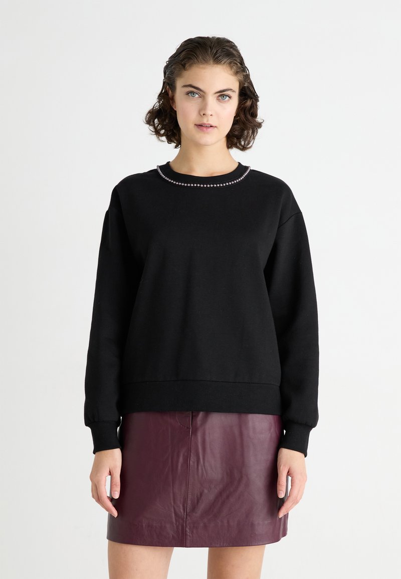 Felpa nera con scollo rotondo, caratterizzata da un dettaglio di perline lungo il colletto. Il modello la abbina a una gonna in pelle bordeaux.