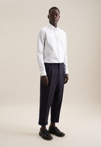 Homme portant une chemise blanche à manches longues boutonnée, un pantalon court plissé foncé et des chaussures habillées noires, debout devant un fond clair.
