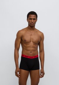 Μαύρα ανδρικά εσώρουχα boxer με κόκκινη ζώνη μέσης με το λογότυπο HUGO, διαθέτουν εφαρμοστή σχεδίαση και λείο, ελαστικό ύφασμα. Το μοντέλο στέκεται ίσια μπροστά σε φωτεινό φόντο.
