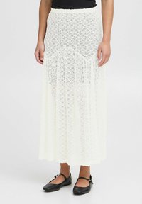 Jupe maxi en dentelle blanche avec un design fluide. Présente une taille festonnée et un ourlet multiple, portée avec des chaussures plates noires.