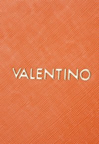 Orange strukturierter Hintergrund mit erhabenem Muster; das goldene "VALENTINO"-Logo prominent platziert.