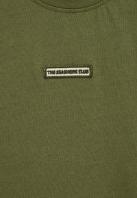 Tissu vert olive avec un petit patch rectangulaire portant l'inscription "THE SEASHORE CLUB" en lettres foncées, centré près du haut.