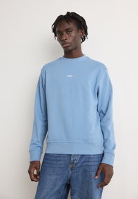 Ανοιχτό μπλε φούτερ με ριμπ ύφανση στους καρπούς και στο τελείωμα, διαθέτοντας ένα μικρό λευκό λογότυπο "BOSS" κεντραρισμένο στο στήθος. Φοράει με μπλε τζην παντελόνι.