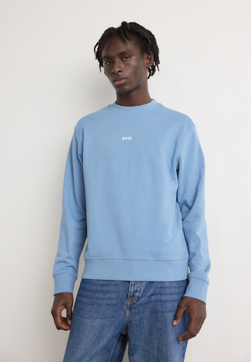 Ανοιχτό μπλε φούτερ με ριμπ ύφανση στους καρπούς και στο τελείωμα, διαθέτοντας ένα μικρό λευκό λογότυπο "BOSS" κεντραρισμένο στο στήθος. Φοράει με μπλε τζην παντ�ελόνι.