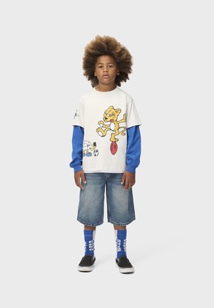 Barn med stort krøllet hår iført lagdelt hvid og blå langærmet skjorte med tegneserie-løve, denimshorts, blå sokker og sorte slip-on sko.