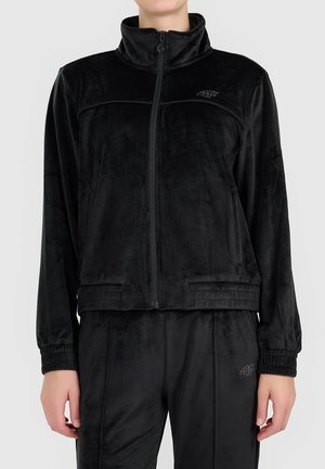 Personne portant une veste polaire noire zippée et un pantalon assorti avec des poignets élastiques et des logos brodés discrets sur la poitrine et la cuisse.