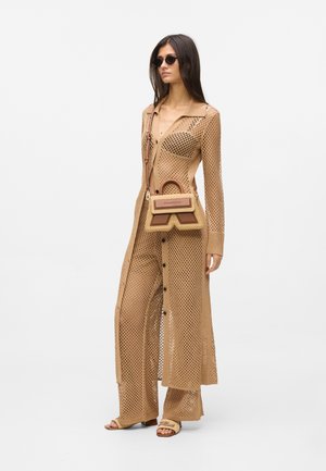 Femme portant un long manteau en maille ouverte couleur beige et un pantalon assorti, tenant un petit sac à main beige et marron, portant des sandales et des lunettes de soleil.