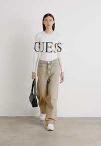 Haut à manches longues blanc avec du texte noir "GUESS", associé à un jean beige clair décontracté et des baskets blanches, tenant un petit sac à main noir.