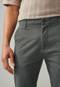 Pantalon gris en tissu lisse avec fermeture à bouton standard et poches latérales, présentant un design à jambes droites et des coutures minimales.